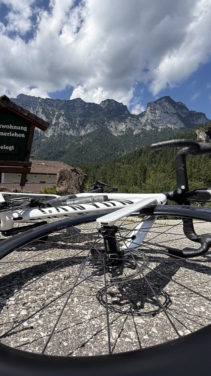 Close-up of a white road bike's front wheel, mountains and clouds behind